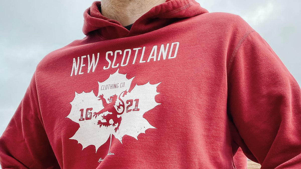 Red hoodie with 'New Scotland' and lion emblem design on a person sitting outdoors.