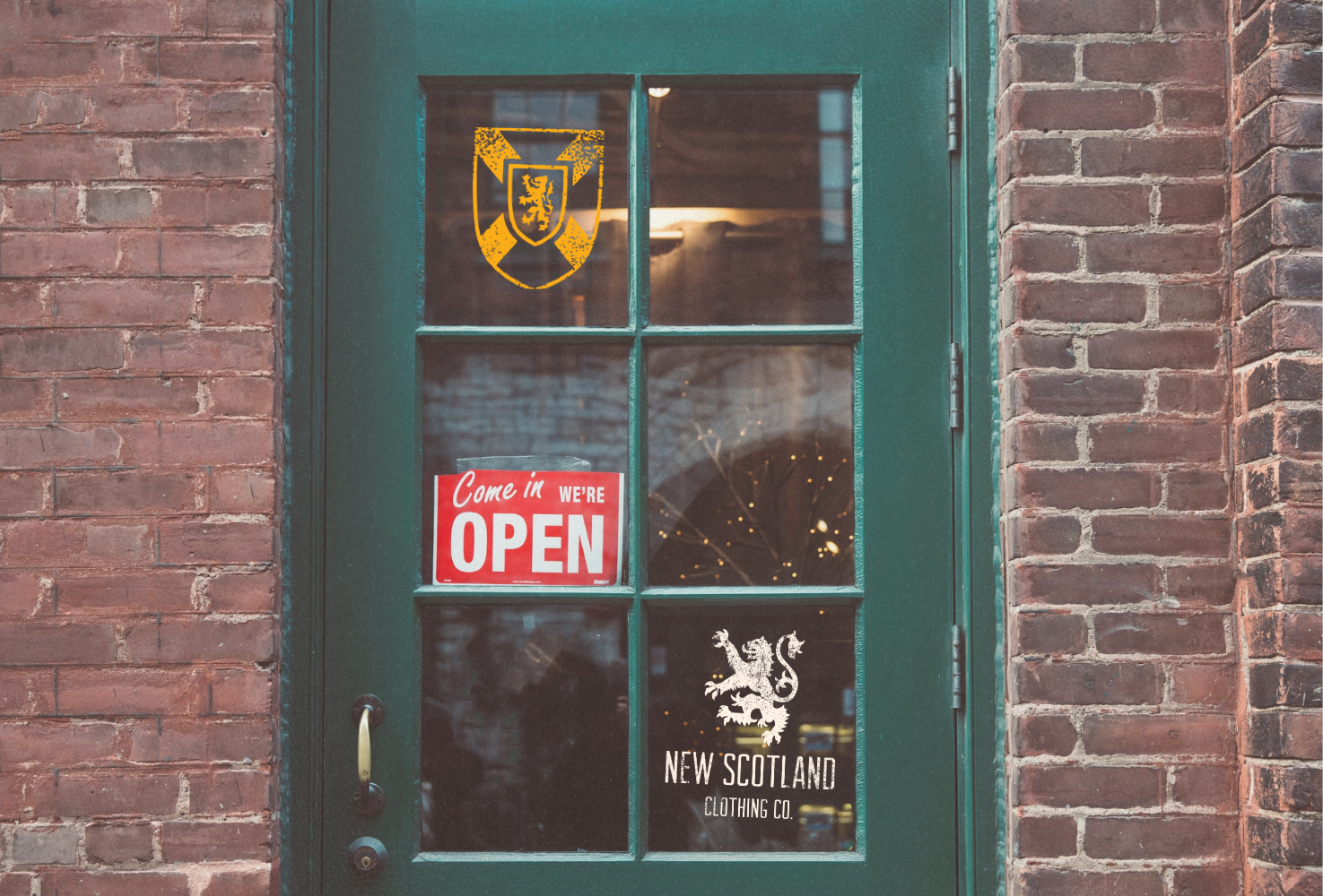 Green door with glass panels featuring a 'New Scotland Clothing Co.' logo and an 'Open' sign, against a brick wall.